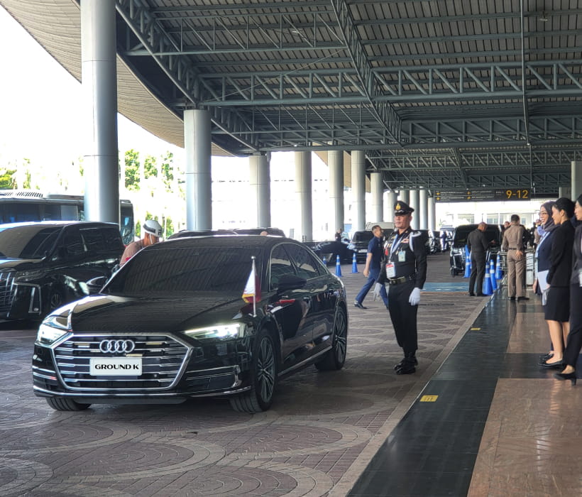 Luxury sedans with flagpoles waiting at the airport exit with military and police standing nearby, surrounded by aides.